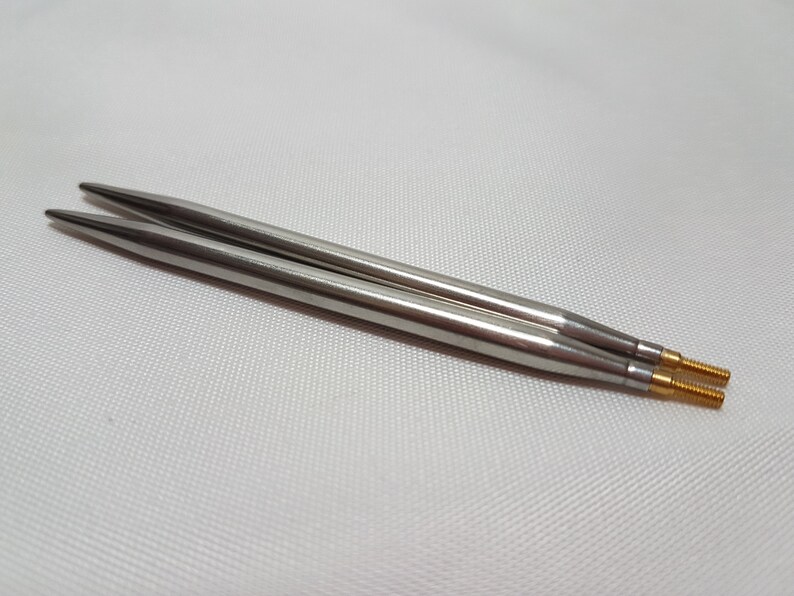 Puede incluir: Dos agujas de tejer plateadas con puntas doradas. Las agujas est&aacute;n sobre una superficie blanca.