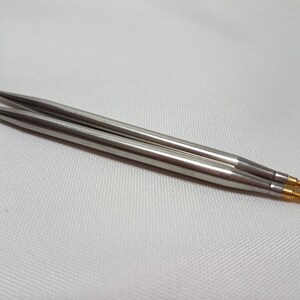 Puede incluir: Un par de agujas de tejer plateadas con puntas doradas. Las agujas est&aacute;n sobre una superficie blanca.