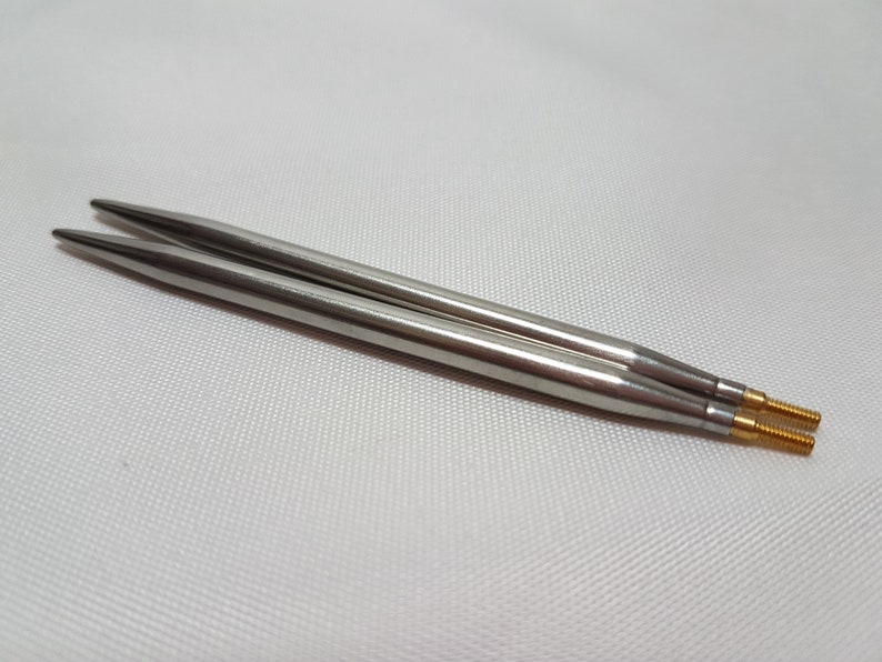 Puede incluir: Un par de agujas de tejer plateadas con puntas doradas. Las agujas est&aacute;n sobre una superficie blanca.