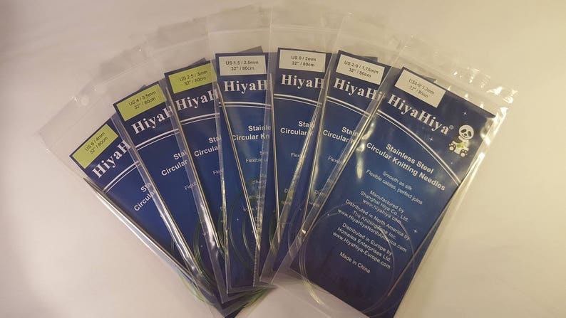 May include: A set of eight HiyaHiya stainless steel circular knitting needles in various sizes. The needles are packaged in clear plastic sleeves with blue and white labels. The labels include the size of the needles in US and metric measurements, as well as the brand name HiyaHiya.