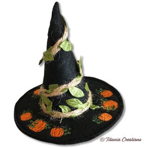 May include: A black felt witch hat featuring embroidered orange pumpkins around the brim. The cone is wrapped with natural jute twine and artificial green leaves. The text Titania Creations is printed in the bottom right corner.