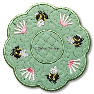 May include: A green, flower-shaped coaster with embroidered details. The coaster features a quilted pattern, with embroidered bees and daisies. The centre has a circular area for a cup. The text "Titania Creations" is visible. The coaster is approximately 13 cm in diameter.