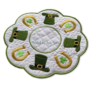 May include: White quilted coaster with a scalloped edge, featuring green leprechaun hats and gold horseshoes with shamrocks. The center has a circular quilted design. Text reads "Titania Creations" and "Stitched by Aida Grier."