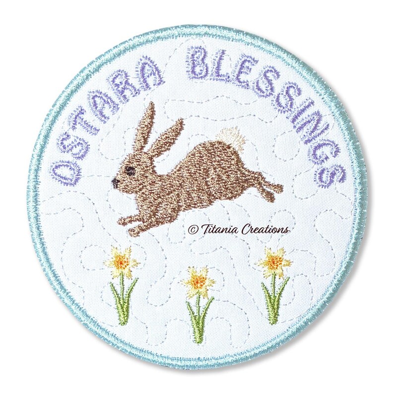 Może przedstawiać: Haftowana naszywka z napisem "OSTARA BLESSINGS" w jasnofioletowym kolorze. Brązowy kr&oacute;lik skacze po białej tkaninie, z trzema ż&oacute;łtymi żonkilami na dole. Naszywka ma jasnoniebieską obw&oacute;dkę i napis "&copy; Titania Creations".