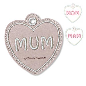 May include: A heart-shaped tag made of light pink material with white embroidered stitching and the word MUM in white thread. The tag features a circular hole at the top for hanging and the text Titania Creations printed below the word MUM.