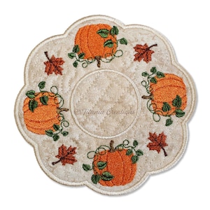 Puede incluir: Un posavasos acolchado redondo con bordes festoneados en tela color crema, con un diseño bordado de cuatro calabazas naranjas con enredaderas verdes y hojas de otoño marrones. El centro tiene un patrón acolchado y el texto Titania Creations.