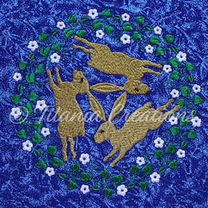 May include: Three brown embroidered rabbits are jumping in a circle surrounded by green leaves and white flowers on a blue background.