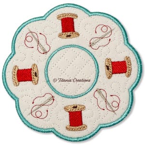 Puede incluir: Un posavasos blanco, acolchado, con forma de flor y borde turquesa. El posavasos presenta carretes de hilo rojo bordados y agujas con hilo. El centro tiene un círculo acolchado. El texto "Titania Creations" es visible.
