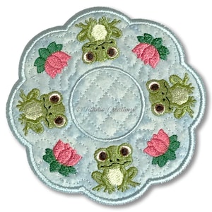 Op de afbeelding: Een lichtblauwe onderzetter met geschulpte rand en een gewatteerd patroon. De onderzetter heeft geborduurde groene kikkers en roze bloemen langs de rand. Het midden heeft een cirkelvormig ontwerp. De tekst "Titania Creations" is zichtbaar.
