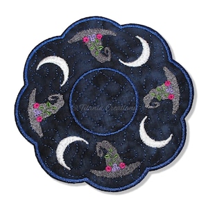 May include: A circular, scalloped-edge fabric coaster in dark navy blue with a quilted texture. It features an embroidered pattern of three silver witch hats adorned with pink and purple flowers, alternating with three white crescent moons.