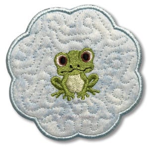 May include: Embroidered frog coaster with a light blue background and a scalloped edge. The frog is green with red-brown eyes and a white belly. The coaster has a quilted pattern and the text "Tanya Creations".