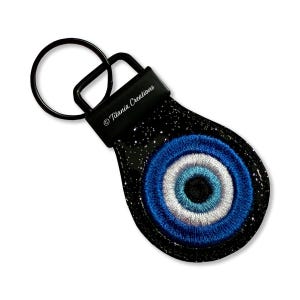 May include: Black keychain with a stitched evil eye design. The eye features concentric circles of blue, white, and a darker blue center. The keychain has a black ring and a strap with the text "Titania Creations".