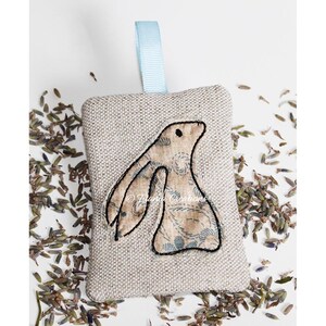 May include: A beige linen sachet with a blue ribbon hanger and an embroidered brown bunny with a blue floral pattern. The sachet is filled with dried lavender buds.