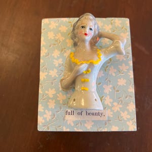 Puede incluir: Figura de cerámica de una mujer con cabello gris, labios rojos y cuello amarillo, montada sobre un fondo azul claro con motivos florales blancos. El texto "full of beauty" está debajo.