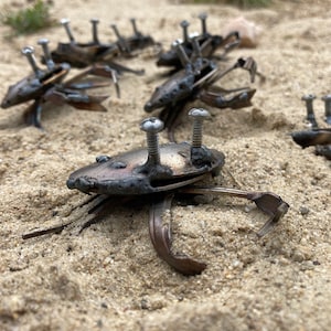 May include: A collection of metal crabs crafted from recycled materials. The crabs are positioned in a line on a sandy surface. Each crab is unique in size and detail, but they share a similar design. These crabs are a creative and unusual way to reuse metal.