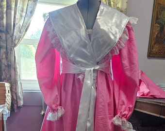May include: A pink satin robe with a white satin collar and lace trim. The robe has a white satin sash and is tied at the waist.