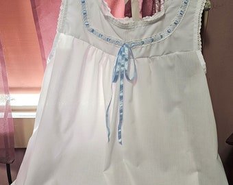 May include: White cotton nightgown with a blue ribbon bow and lace trim around the neckline. The nightgown has a ruffled hem.