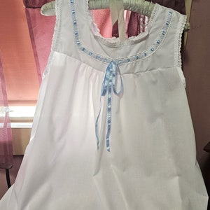 Puede incluir: Camisón blanco de algodón con un lazo de cinta azul y ribete de encaje alrededor del escote. El camisón tiene un dobladillo con volantes.