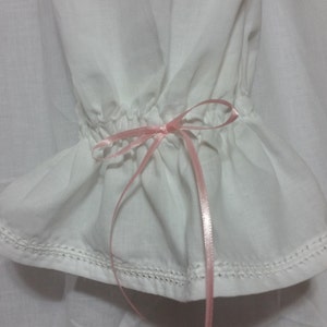 May include: White cotton fabric with a pink ribbon tied in a bow at the end of the fabric. The fabric has a ruffled edge and is sewn with a white thread.