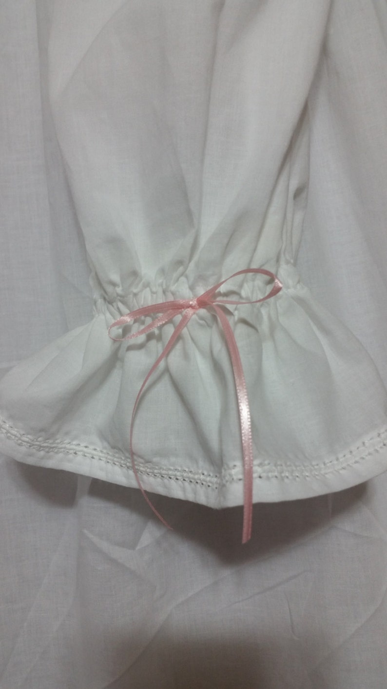 May include: White cotton fabric with a pink ribbon tied in a bow at the end of the fabric. The fabric has a ruffled edge and is sewn with a white thread.