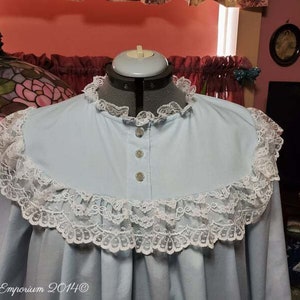 Puede incluir: Un camis&oacute;n azul claro con un ribete de encaje blanco alrededor del cuello y las mangas. El camis&oacute;n tiene un frente abotonado.
