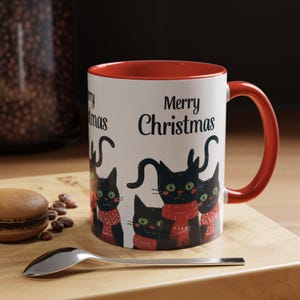 Peut inclure: Mug en céramique blanche avec intérieur et anse rouges. Le mug présente des illustrations de chats noirs portant des écharpes rouges, avec les mots "Merry Christmas" imprimés en noir. Une cuillère et un macaron sont sur une surface en bois.