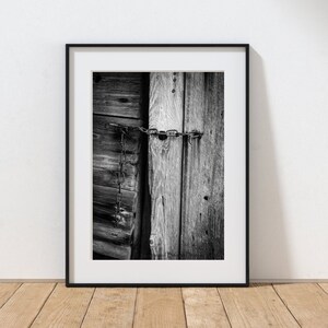 May include: Black and white photograph of a wooden door with a chain and lock. The door is made of weathered wood planks and has a rustic look. The chain is wrapped around the door and is secured with a padlock.