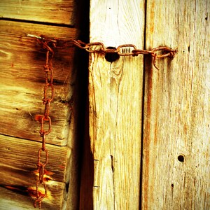 May include: A close-up of a wooden door secured with a rusty chain. The wood is weathered and has a warm, golden hue. The chain is wrapped around the door and secured with a padlock.