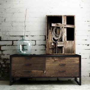 May include: A rustic wooden dresser with three drawers and black metal legs. A clear glass vase with a twig sits on top of the dresser. A framed black and white photograph of a wooden crate with rope and wood inside hangs on a white brick wall behind the dresser.