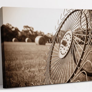 May include: Sepia-toned canvas print of a hay rake in a field. The rake's metal wheel is in sharp focus, with curved tines and a central hub. In the background, blurred hay bales and trees are visible, evoking a rural, agricultural theme.