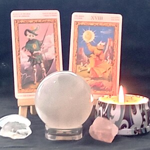 May include: A crystal ball on a clear stand, tarot cards, a lit candle, and crystals are arranged on a dark surface. The tarot cards feature illustrations with gold frames. A pink crystal and a clear crystal are also present.
