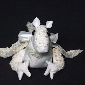 May include: A white stuffed animal with a bow, decorative embellishments, and black eyes. The plush toy has white gloves and is set against a black background. The toy's wings are decorated with beads.
