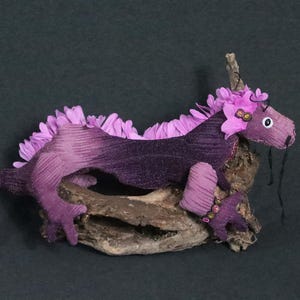 May include: A handmade purple dragon plush toy with a flower mane and accents. The dragon is made of fabric and sits on a piece of driftwood. The dragon's body is a gradient of purple hues.