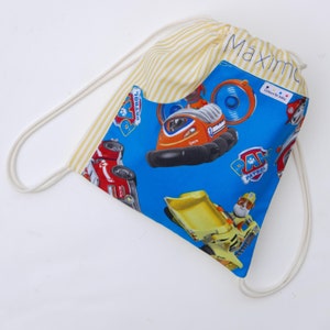 May include: A blue drawstring bag with a yellow and white striped pocket. The bag features a Paw Patrol theme with images of the characters' vehicles and the words "Paw Patrol". The bag has a white drawstring and the name "Maximo" is written on the pocket.