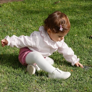May include: A young child wearing a pink and white polka dot blouse with a ruffled collar and white tights sits on a grassy lawn. The child is wearing white shoes with a buckle.
