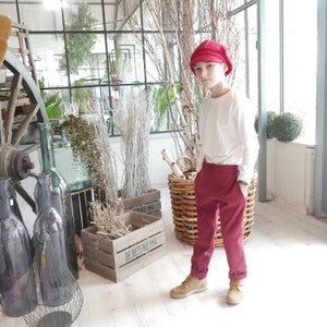 May include: A young person wearing a red newsboy cap, a white long-sleeved shirt, and red striped pants. The person is standing in a room with a wooden crate and a wicker basket.