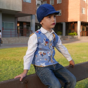 Puede incluir: Un ni&ntilde;o peque&ntilde;o con un chaleco estampado en azul y blanco con una gorra azul. El chaleco tiene un tema de vaquero con estrellas e im&aacute;genes de vaqueros. El ni&ntilde;o est&aacute; sentado en un banco de madera.
