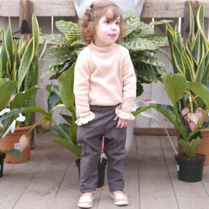 May include: A young girl wearing a light pink sweater and dark gray pants with a white pattern. She is standing in front of a wall with potted plants.