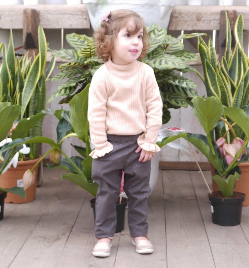 May include: A young girl wearing a light pink sweater and dark gray pants with a white pattern. She is standing in front of a wall with potted plants.