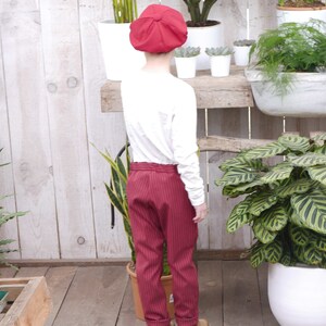 May include: A child wearing a red newsboy cap, a white long-sleeved shirt, and red striped pants. The child is standing in front of a wooden shelf with plants.