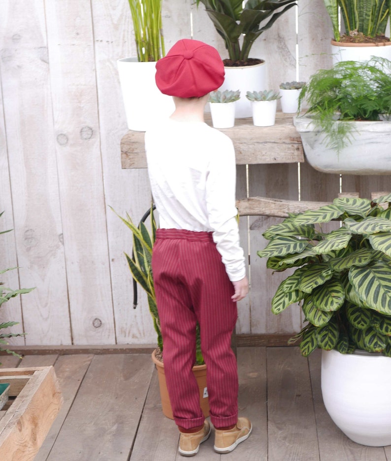May include: A child wearing a red newsboy cap, a white long-sleeved shirt, and red striped pants. The child is standing in front of a wooden shelf with plants.