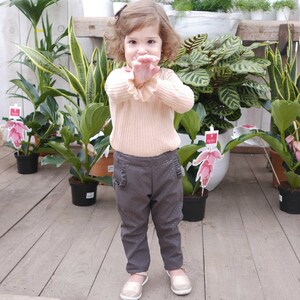 May include: A young girl wearing a light pink sweater and dark gray pants with white polka dots. She is standing in a greenhouse with plants in the background.