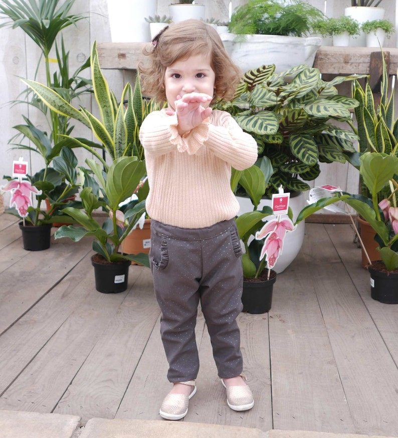 May include: A young girl wearing a light pink sweater and dark gray pants with white polka dots. She is standing in a greenhouse with plants in the background.