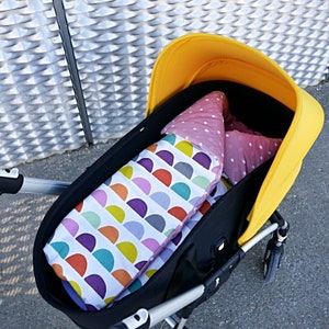 K&ouml;nnte beinhalten: Ein gelber Kinderwagen mit schwarzer Stoffauskleidung und einer bunten, gemusterten Decke im Inneren. Die Decke hat ein geometrisches Muster mit Kreisen und Halbkreisen in den Farben Lila, Gr&uuml;n, Gelb und Rosa.