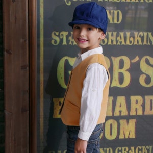 Puede incluir: Una persona joven con una gorra de repartidor de peri&oacute;dicos azul, una camisa blanca abotonada, un chaleco amarillo y pantalones vaqueros azules. La persona est&aacute; de pie frente a un letrero que dice "G.B.'s Billiard Room".