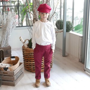 May include: A young person wearing a white long-sleeved shirt, red striped pants, and a red newsboy cap. The person is standing in front of a wicker basket.