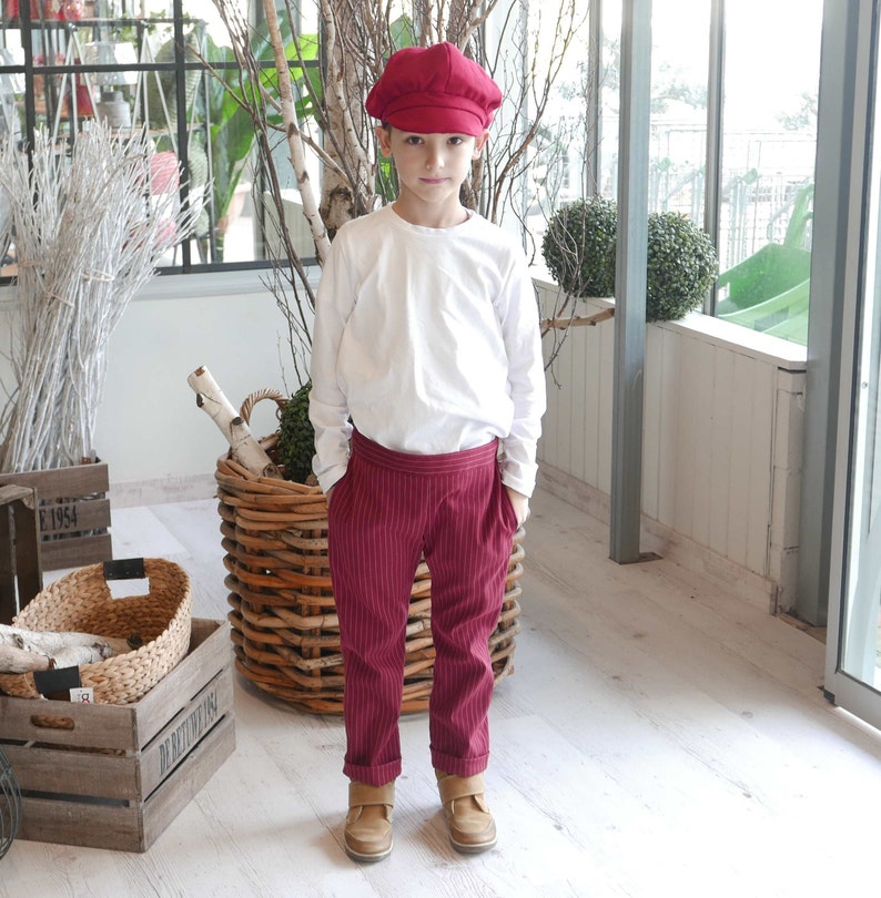 May include: A young person wearing a white long-sleeved shirt, red striped pants, and a red newsboy cap. The person is standing in front of a wicker basket.