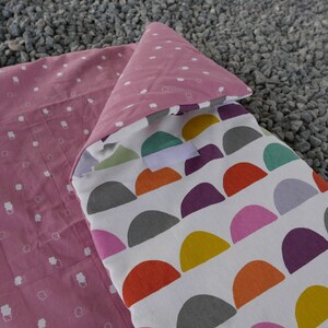 K&ouml;nnte beinhalten: Ein rosa-wei&szlig;er Schlafsack mit einem wei&szlig;en Stofffutter, das ein farbenfrohes geometrisches Muster aus Halbkreisen aufweist.