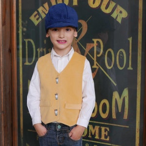 Puede incluir: Una persona joven que lleva un chaleco amarillo con botones de estampado de cebra, una camisa blanca, pantalones vaqueros azules y una gorra de newsboy azul. La persona est&aacute; de pie frente a un cartel que dice "Enjoy Our Drink & Pool Room".
