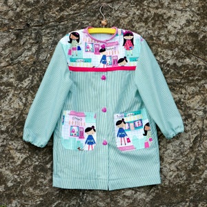 May include: A light blue and white striped smock with a pink trim and colorful cartoon girls on the front. The smock has two pockets with a cartoon girl on each pocket. The smock has a button closure.
